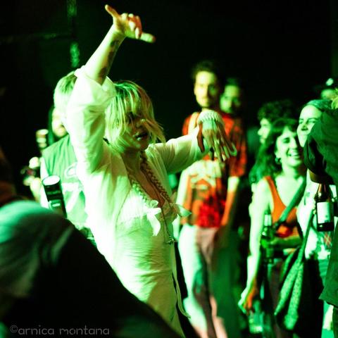 A person with blonde hair is dancing energetically in a lively crowd, illuminated by green lights. Surrounding them are various festival-goers enjoying the performance, some holding drinks and smiling. The atmosphere is vibrant and filled with excitement.