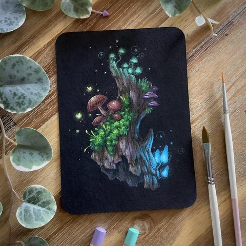 A photo of a small painting on black paper with rounded corners, placed on a wooden table and surrounded by coloured pencils, paint brushes, and leaves for decoration. The painting features a seemingly floating log overgrown with moss and lots of different mushrooms, some luminescent. Tiny fairies, mere orbs of light, hover around the mushroom.