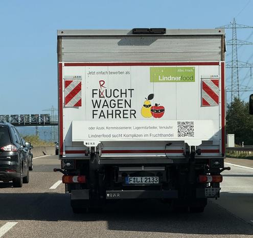Das Bild zeigt die Rückseite eines weißen Lastwagens, der auf einer Autobahn fährt. Im Zentrum der LKW-Rückseite befindet sich eine große Werbefläche. Dort steht in großen schwarzen Großbuchstaben: „FLUCHTWAGENFAHRER“, wobei das Wort kreativ verändert wurde. Das “L” in „FLUCHT“ ist durch ein rotes „R“ überklebt, sodass man sowohl „FRUCHTWAGENFAHRER“ als auch „FLUCHTWAGENFAHRER“ lesen kann – ein Wortspiel mit Humor.

Neben dem Text sind zwei gezeichnete Früchte abgebildet:
	•	eine Birne mit Sonnenbrille (die wie eine Maske aussieht),
	•	ein Apfel mit schwarzer Augenmaske.

Beide Früchte sehen aus wie kleine Gangster oder „Komplizen“. Darüber steht in kleiner Schrift:
„Jetzt einfach bewerben als“
und darunter:
„oder Azubi, Kommissionierer, Lagermitarbeiter, Verkäufer“

Darunter in einer weißen Leiste steht in kleinerer Schrift:
„Lindnerfood sucht Komplizen im Fruchthandel!“

Rechts oben ist das Logo von Lindnerfood mit dem Slogan „Alles. Frisch.“ abgebildet. Rechts unten ist ein QR-Code zum Scannen.