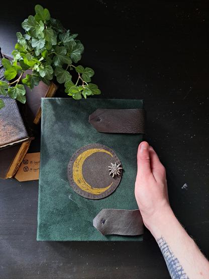 An a5 sized dark green handmade sketchbook with brown leather clasps and a brown leather patch in the middle. The leather patch has a golden sicle moon and an edelweiss flower button on it.