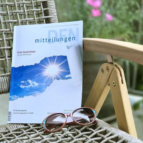 Ein Exemplar der Zeitschrift „DFN-Mitteilungen“ liegt auf einem Gartenstuhl mit geflochtener Sitzfläche. Der Titel der Ausgabe lautet „Gute Aussichten mit der DFN-Cloud“ und zeigt eine Sonne, die durch eine Wolkenlücke strahlt. Vor dem Heft liegt eine rot-türkis gerahmte Sonnenbrille. Im Hintergrund sind unscharf grüne Pflanzen und pinke Blüten zu sehen – sommerliche Atmosphäre im Freien. Foto: DFN