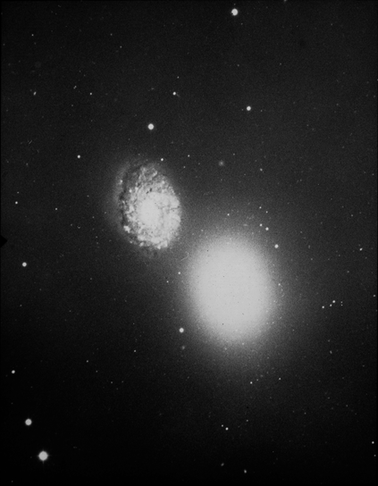 A grainy, black and white image of a large elliptical galaxy and a smaller face-on spiral galaxy. The elliptical galaxy is in the lower right portion of the frame. It appears as a featureless, oval-shaped blob surrounded by a haze of stars. Above and to its left is its companion, a smaller face-on spiral galaxy. Its somewhat poorly defined spiral arms curve around its bright center. The arms are dotted with bright stars and star clusters and are crossed by a series of filamentary dust lanes. Many white dots, which are stars, are seen on the black background.
