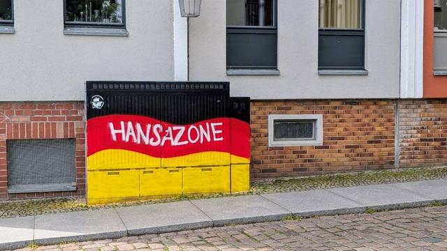 Vor einem Haus steht ein vollständig besprühter Stromkasten. Der Untergrund besteht aus drei waagerechten Flächen in schwarz/rot/gelb wie die deutsche Nationalflagge. Die Farbflächen sind dabei wellig gestaltet, um eine wehende Fahne zu symbolisieren.

Oben links im schwarzen Bereich wurde ein kleines weißes FC Hansa-Logo platziert.

In der Mitte zieht sich im roten Bereich in großen Buchstaben der Schriftzug „HANSAZONE“ von links nach rechts.