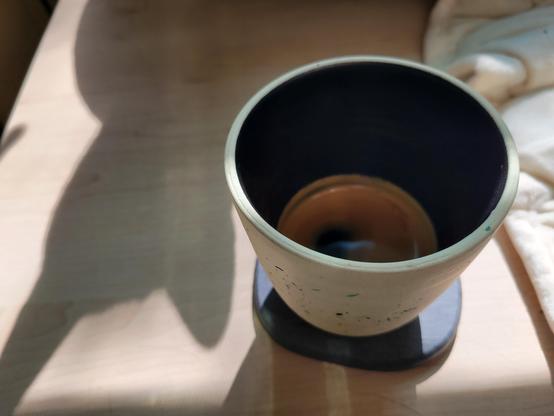 12-ounce, handle-less, off-white ceramic cup with a coffee pumpkin-spice latte, on a gray diatomaceous-earth coaster on a maple-veneer desk. The clay of the cup has many tiny flecks of black and blue. The interior of the cup has a dark-colored glaze.

The early-morning sun is bright through a window. A shadow of a cat face is seen on the desk to the left.