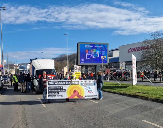 Das Bild zeigt eine Demonstration auf einer Straße bei sonnigem Wetter. Im Vordergrund halten zwei Personen ein Banner mit der Aufschrift:
„Nie wieder ist jetzt! Demokratie & Menschenwürde sind nicht verhandelbar!“
Darunter ist ein Symbol mit zwei sich haltenden Händen vor einem gelben Hintergrund zu sehen. Dahinter steht ein weißer Transporter mit weiteren Menschen. Rechts im Bild befinden sich ein CinemaxX-Kino sowie ein großer digitaler Werbebildschirm mit einer 1&1-Werbung.

Im Hintergrund sieht man viele weitere Demonstrierende und ein großes Polizeiaufgebot, das die Straße sichert. Insgesamt vermittelt das Bild den Eindruck einer friedlichen, gut besuchten Kundgebung für Demokratie und Menschenrechte.