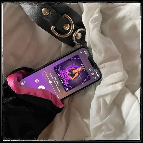 On white sheets, an iPhone playing Kiss of Seduction by Rawnie Sabor, narrated by Allie Shae, coming out of a black velvet pouch, with pink satin lining. Next to the phone is a leather handcuff with silver buckles.
