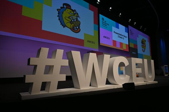 #WCEU hashtag as physical letters on stage at this WordCamp Europe 2025 in Basel, with the special-edition Wapuu (WP mascot) on screen in the background.