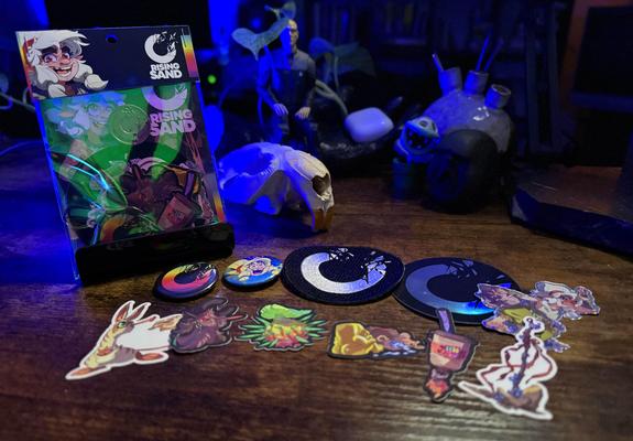 Photo of Rising Sand merchandise; several stickers of characters and items from the comic, patches, and buttons. There's a sealed package of the same items with Rising Sand branding.