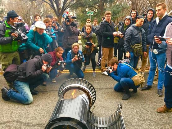 A gaggle of journalists and photographers cynically gather around a single, overturned trash bin, the contents of which are slightly on fire, in order to turn it into a dramatic "violence" narrative at a protest.