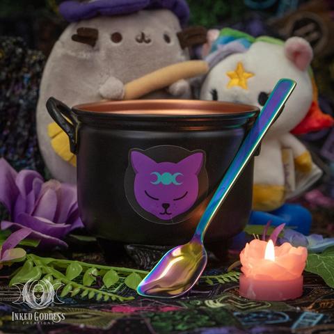 A small black Wee Witches Metal Cauldron and Spoon Set sits in the center of the image. The cauldron has two handles, one on either side, a stylized cat head with triplemoon decal on the front, and a copper-colored inside surface. It has a rainbow-colored metal spoon leaning against it. This is an exclusive product from Inked Goddess Creations.