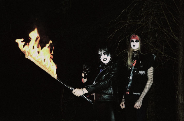 the duo wearing leather and corpse paint, one holding a flaming sword, in a darkened wood