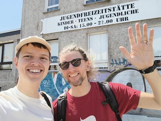 Vor dem Gebäude der Jugendfreizeitstätte in Lütgendortmund stehen zwei lächelnde Männer im Sonnenlicht. Rechts im Bild: Benjamin Beckmann mit Sonnenbrille, rotem T-Shirt und Rucksack, der in die Kamera winkt. Links daneben ein Fraktionskollege mit heller Kappe. Über ihnen prangt ein Schild mit der Aufschrift: „JUGENDFREIZEITSTÄTTE – KINDER · TEENS · JUGENDLICHE – DI–SA: 15.00–21.00 UHR“. Die Stimmung ist freundlich und offen, im Hintergrund ist Graffiti sichtbar.