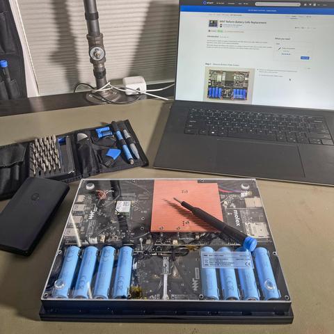 MNT Reform laptop flipped over on a workbench. The laptop's translucent bottom cover reveals its internals, which notably features blue 18650 batteries instead of a proprietary one. Sitting on top of the laptop is an iFixit screwdriver, and in the background is an open Pro Tech toolkit and a laptop with the MNT Reform battery replacement guide open on iFixit.com.