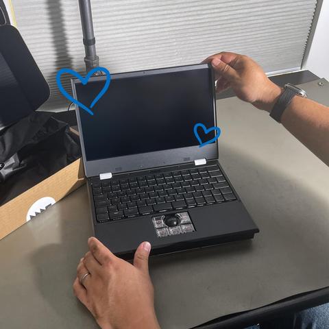 MNT Reform laptop sitting on a workbench/desk. Two hands are touching the laptop, one holding its base while the other opens its lid. The laptop's shipping box is sitting to its left, implying it was just opened.