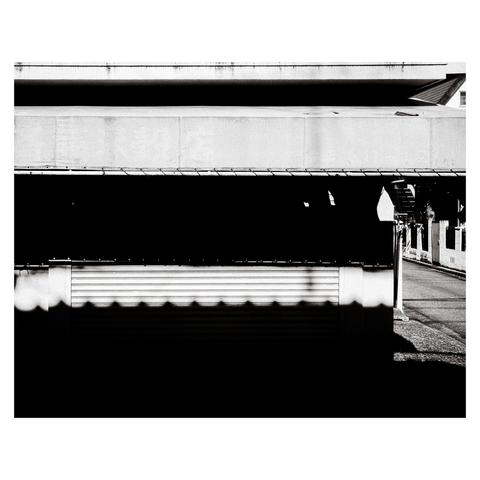 This black-and-white photo captures the sharp contrast of midday shadows on a closed shopfront in Nagoya. The lower half of the image is nearly black, with only the faint silhouette of a metal shutter visible. A scalloped roof shadow falls across the shutter, echoing the shape of traditional Japanese roof tiles. Above it, a tattered awning juts out, casting an irregular shadow that merges into the larger dark expanse.