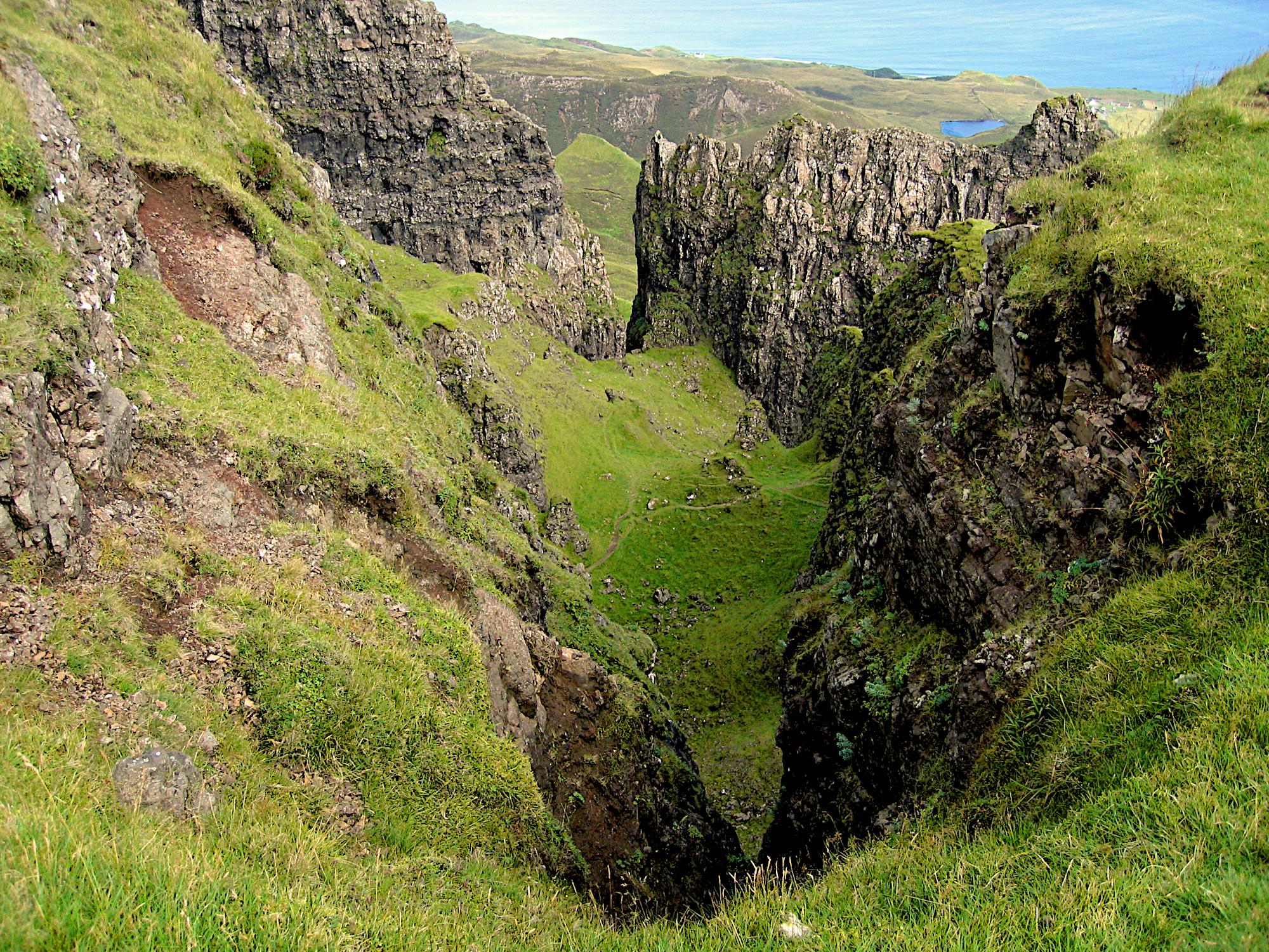 Das Bild zeigt eine Felsenschlucht am Berg Quiraing auf der Insel Isle of Skye. Eine flache Schlucht zwischen groben Felswänden, alle geraden Flächen sind mit Gras bewachsen und grün. Ganz am oberen Rand ist im Hintergrund das Meer zu erkennen. Das Bild zeigt eine Felsenschlucht am Berg Quiraing auf der Insel Isle of Skye. Eine flache Schlucht zwischen groben Felswänden, alle geraden Flächen sind mit Gras bewachsen und grün. Ganz am oberen Rand ist im Hintergrund das Meer zu erkennen.