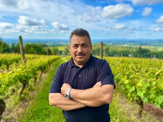 Moises Sotelo, arms crossed, looks into the camera. He is standing in a vineyard on a sunny day.
