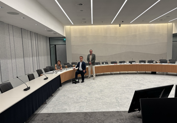 Foto van een lege Commissiezaal in de Tweede Kamer: een grijze zaal met een halfronde cirkel met tafels. Links in een hoek zit in, aan de andere kant van de tafel staan Thibault en Thijs. Foto is wat van een afstand genomen zodat de leegte goed in beeld is. 