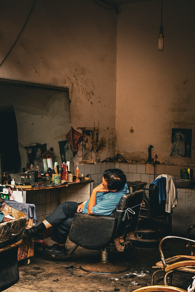 Ein Mann schläft in einem Friseurstuhl in einem einfachen, leicht abgelebten Friseursalon in China. Er sitzt zurückgelehnt, die Augen geschlossen, mitten am Tag. Um ihn herum sind Friseurutensilien und verblasste Spiegel zu sehen. Die Szene wirkt ruhig, fast zeitlos – ein stiller Moment im Trubel des Alltags. 