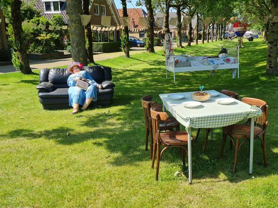 Leren bank met een dikke mevrouwenpop. Op de voorgrond een gedekte tafel met vier stoelen. Op de achtergrond een zwevend bed aan touwen, bevestigd aan de bomen. Het tafereel staat buiten op een smalle grasstrook, omringd door bomen.