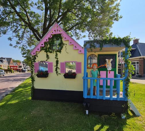 Nagebouwde Villa Kakelbont. Gele gevel, roze luikjes, roze dakrand, planten, blauwe veranda met een als Puppi verklede pop en achter haar een wit paard in een stal met roze staldeur.