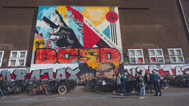 A street photo of a brick wall with a large (several stories tall) painted panel mounted on it. On the panel: a black and white graphics of a big V sign (✌), some geometric shapes in red and yellow, words "O B E Y  In The Name Of Humanity" and blockly letter BGSD at the bottom.
Below the panel, large letters are spray-painted in silver and red, a cartoonish face and a fist are painted between these letters, in the centre of the wall.
Lots of bicycles are parked in front of these letters. Some people are standing next to be bikes on the right side of the image.