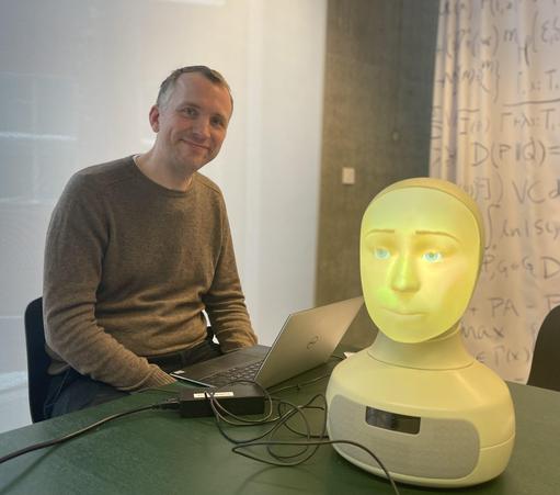 Gabriel sitting at a desk next to a laptop and Furhat robot - a robotic head with a face back-projected inside it. 