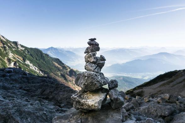 Ein Steinmanndl in einer Felslandschaft in den Bergen. Leicht diesig blickt der Betrachter auf weitere, niedrigere Bergkuppen. Ein sonniger, praktisch wolkenloser Tag, der Betrachterstandort liegt aber im Schatten.