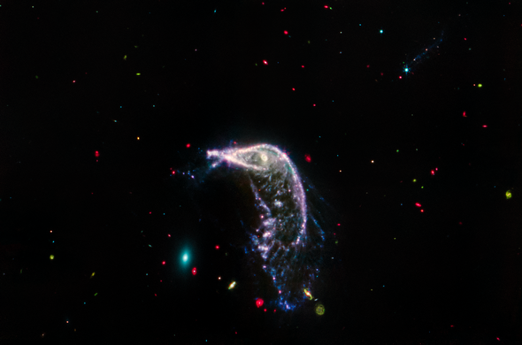 Two interacting galaxies known as Arp 142 in a horizontal image taken in mid-infrared light. At left is NGC 2937, an elliptical galaxy that looks like a tiny teal oval and is nicknamed the Egg. At right is NGC 2936, a distorted spiral galaxy nicknamed the Penguin, which is significantly larger. A beak-like region points toward the Egg, but lies far above it. Where the eye would be is an opaque, almost washed-out pink spiral. This galaxy’s distorted pink, purple, and blue arms create the bird’s beak, back, and tail. The tail, which is closer to the Egg, is wide and layered, like a beta fish’s tail. The Penguin and the Egg appear very separate. The galaxy at top right, PGC 1237172, is barely visible. A brighter slightly larger blue foreground star that is over top of this galaxy has tiny diffraction spikes. Throughout the image are tiny galaxies in bright reds, greens, and blues. The background of space is black.