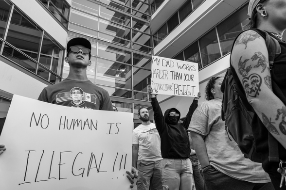 Black and white image of an ICE protest in Des Moines, Iowa. Multiple signs can be seen being held including, No Human is Illegal and My Dad Works Harder Than Your Oink Oink President. 