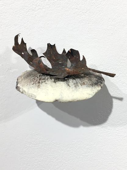 A metal sculpture of a leaf mounted on a white mushroom on a white wall.