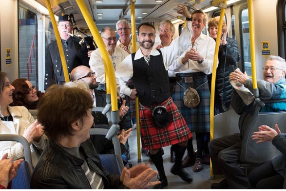 a man in a kilt dancing on a public transit train car. the other passengers are absolutely loving it.