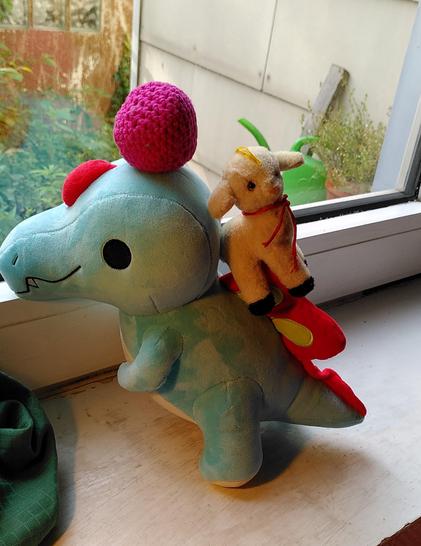 Zilla, die blaue Spinosaurierin,  schaut aus dem Fenster und balanciert dabei einen pinken Ball auf dem Kopf. Derweil rutscht das kleine Plüschschaf Lily ihren schrägen Rücken herab. 