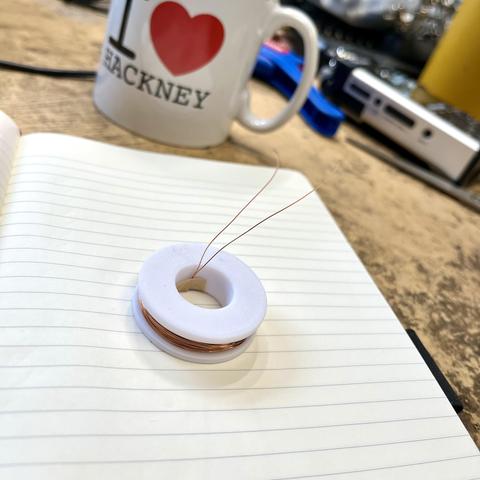 A white plastic 3D-printed bobbin sits on a lined notebook. The bobbin is wound in thin enamelled copper wire and the two ends poke out of it. In the background is my bench with a cheeky I Love Hackney mug crashing the picture.
