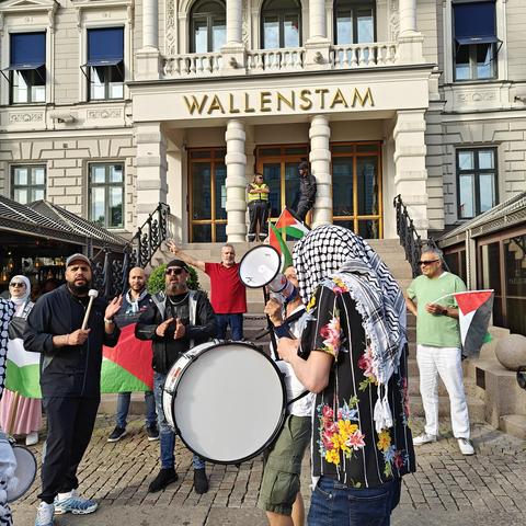 A group of people with kufiyas, flags, and drums, in front of an impressive stone building from the late 19th century