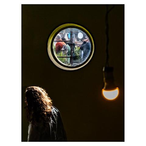 A girl is walking up the stairs in a dark stairwell of a downtown apartment building. Through a small round window we can see the roof, where there are balloons and people. In the right foreground of the picture, a dimly lit incandescent lamp hangs from the ceiling.