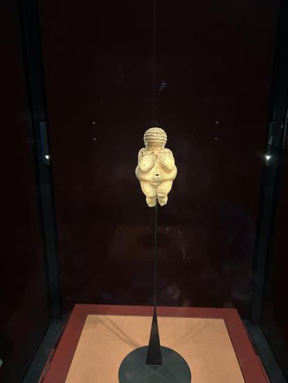 Die Venus von Willendorf in einer Vitrine. Eine ca. 30.000 Jahre alte Figurine aus sandfarbenem Stein, die eine füllige, gesichtslose Frau ohne Kleidung darstellt.