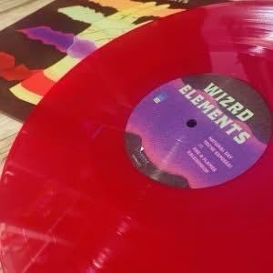 Eine leuchtend rote Vinyl-Schallplatte mit der Aufschrift "WIZRD ELEMENTS" ist abgebildet, neben einem farbenfrohen Album-Artwork im Hintergrund. Das Bild hebt die glänzende Oberfläche und die leuchtenden Farben der Schallplatte hervor.