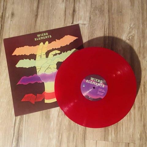 Eine leuchtend rote Vinyl-Schallplatte mit dem Titel "WIZRD ELEMENTS" wird neben ihrem Albumcover ausgestellt, das eine bunte Illustration einer Hand und stilisierte Elemente auf einem dunklen Hintergrund zeigt. Die Szene ist auf einer Holzoberfläche platziert.