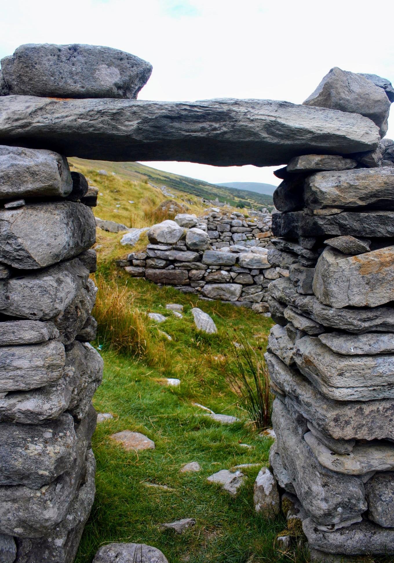 Das Foto zeigt einen Durchgang aus aufgeschichteten grauen Natursteinen, teilweise mit gelben Flechten überzogen 
Der Blick fällt hindurch in die dahinter befindliche Landschaft. Zu sehen ist eine Seite eines Hügels, auf dem sich verteilt weitere dieser Natursteinen befinden, ebenso aufgeschichteten, halbhohe Mauern. Alles wirkt ruhig und verlassen. Das Foto zeigt einen Durchgang aus aufgeschichteten grauen Natursteinen, teilweise mit gelben Flechten überzogen 
Der Blick fällt hindurch in die dahinter befindliche Landschaft. Zu sehen ist eine Seite eines Hügels, auf dem sich verteilt weitere dieser Natursteinen befinden, ebenso aufgeschichteten, halbhohe Mauern. Alles wirkt ruhig und verlassen.