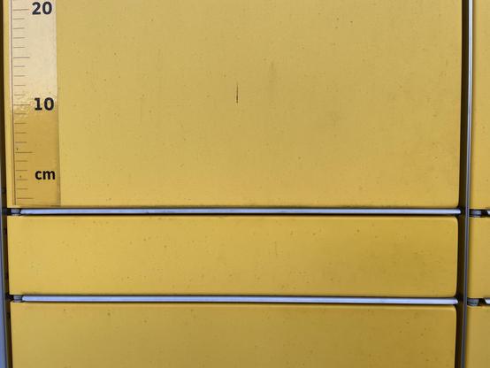 Das Bild zeigt eine Reihe von gelben Schubladen, die in einer Reihe angeordnet sind. Auf der linken Seite des Bildes befindet sich ein Zentimetermaßstab, der von 0 bis 20 cm reicht. Die Schubladen sind gleichmäßig verteilt und durch silberne Streifen getrennt. Die Oberfläche der Schubladen ist glatt und zeigt einige kleine Flecken und Abnutzungserscheinungen. Die Schubladen scheinen aus einem festen Material zu bestehen, möglicherweise Metall oder Kunststoff. Die Farbe der Schubladen ist ein einheitliches Gelb.
