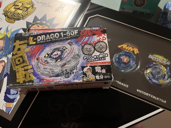 A photo of Beyblades. The focus of the image is an in-box Lightning L-Drago next to a Storm Pegasus from the 25th Anniversary box set. All Beyblades shown are from the Beyblade X line.