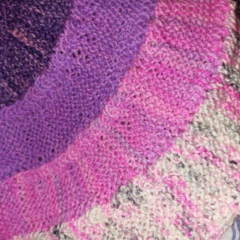 Radiating curves of knitting, from centre out (and top left corner to bottom right) the yarns are- dark purple, purple, pink, and a black and pink speckled white.