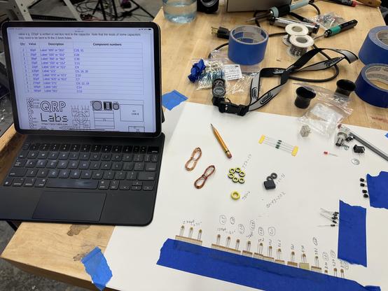 A sheet of paper on a workbench, with various electronic parts taped to it with an ipad next it showing assembly instructions. 