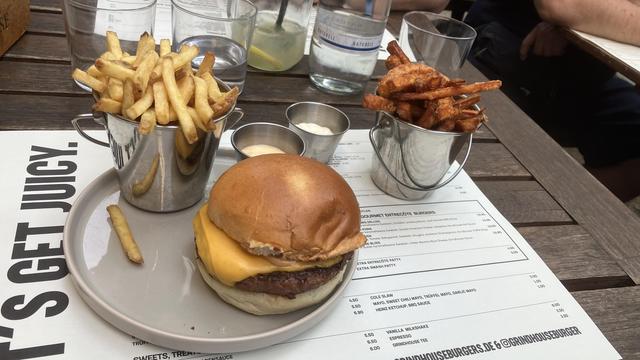 Das Bild zeigt ein Buffet mit einem Burger und Pommes auf einem Holztisch. Der Burger auf einem grauen Teller besteht aus einem bunten Brot, einem Rindfleischpatty und einer Scheibe Käse. Neben dem Burger befinden sich zwei Metallkübel mit Pommes: einer mit goldenen Pommes und einer mit süßen Pommes. Auf dem Teller liegen auch ein paar einzelne Pommes. Zwei kleine Metallbecher enthalten eine weiße Sauce, wahrscheinlich eine Mayonnaise oder eine Ranch-Dressing. Im Hintergrund ist ein Menü mit Preisen und Beschreibungen zu sehen, sowie ein Glas Wasser und ein Glas mit Limettenscheiben. Der Text auf dem Menü lautet "LET'S GET JUICY." und die Website "GRINDHOUSEBURGERS.DE" ist ebenfalls sichtbar.