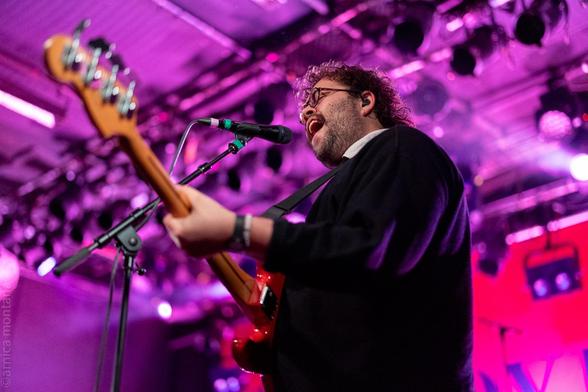 A musician performs on stage, singing into a microphone while playing a red electric bass guitar. The background features colorful stage lighting, primarily in purple tones, and blurred stage elements.