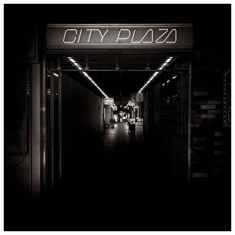 A black and white photograph of a narrow, dimly lit indoor shopping passage labeled "CITY PLAZA." The neon sign above the entrance glows brightly, contrasting with the deep shadows within. Inside the corridor, a line of small illuminated signs and shop boards fades into the darkness, creating a tunnel effect. The scene is almost deserted, lending an eerie, cinematic feel reminiscent of a vintage film noir or a slightly unsettling memory of a retro arcade strip. The lighting is minimal but deliberate, with overhead fluorescent fixtures casting a faint path into the depths of the hallway.