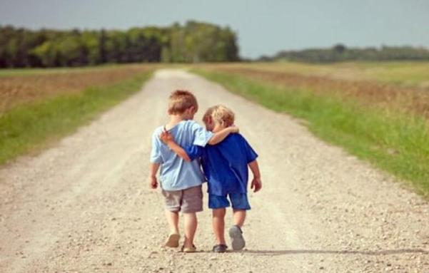The image depicts two young children walking hand in hand down a gravel road. The child on the left is wearing a light blue shirt and gray shorts, while the child on the right is dressed in a blue shirt and blue shorts. They are walking away from the camera, with the child in blue having their arm around the other child's shoulder, indicating a close bond. The road is flanked by green grass and fields on either side, with a line of trees visible in the background. The sky is clear and blue, sug…