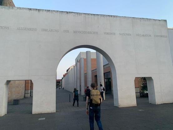 Monument to the Universal Declaration of Human Rights. A row of columns, each column is a different right, written out in various languages. The start is a large wall with 2 doorways on each side of a central arch. The wall and the arches are all white.