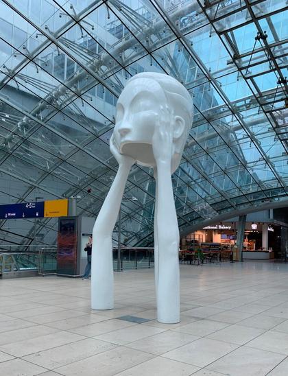 Scuplture in Frankfurt train station, two slender arms holding up a head that is missing a jaw. The whole sculpture is white, with the station metal and glass arches above.
